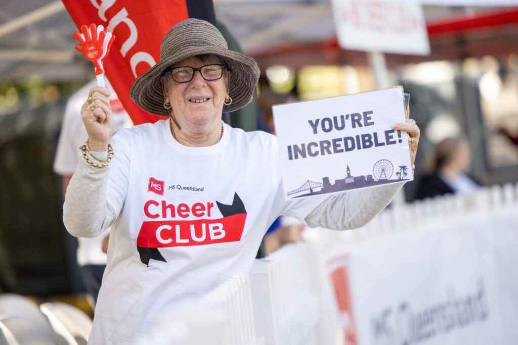 A woman wearing a hat and glasses smiles while holding a sign that says "YOU'RE INCREDIBLE!" and a red clapper toy. She wears an "MS Queensland Cheer CLUB" shirt at an outdoor event.