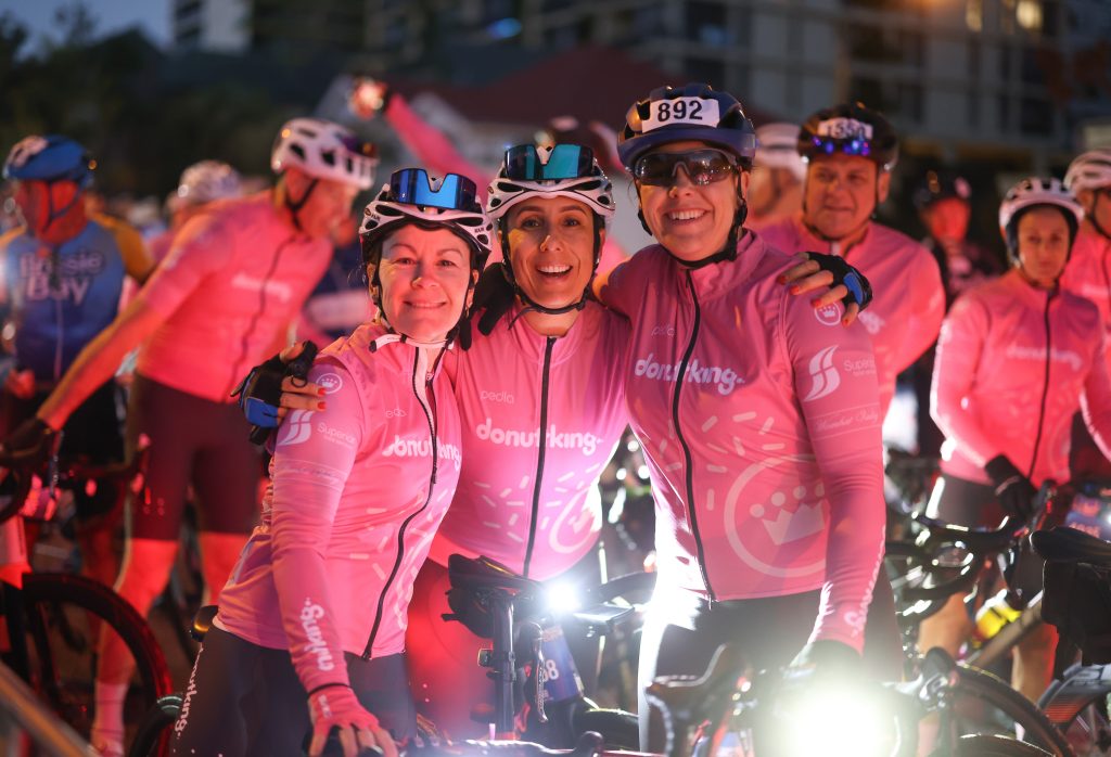 Three smiling cyclists in matching pink jerseys and helmets pose together with their arms around each other at a nighttime cycling event, with other cyclists in similar outfits in the background.