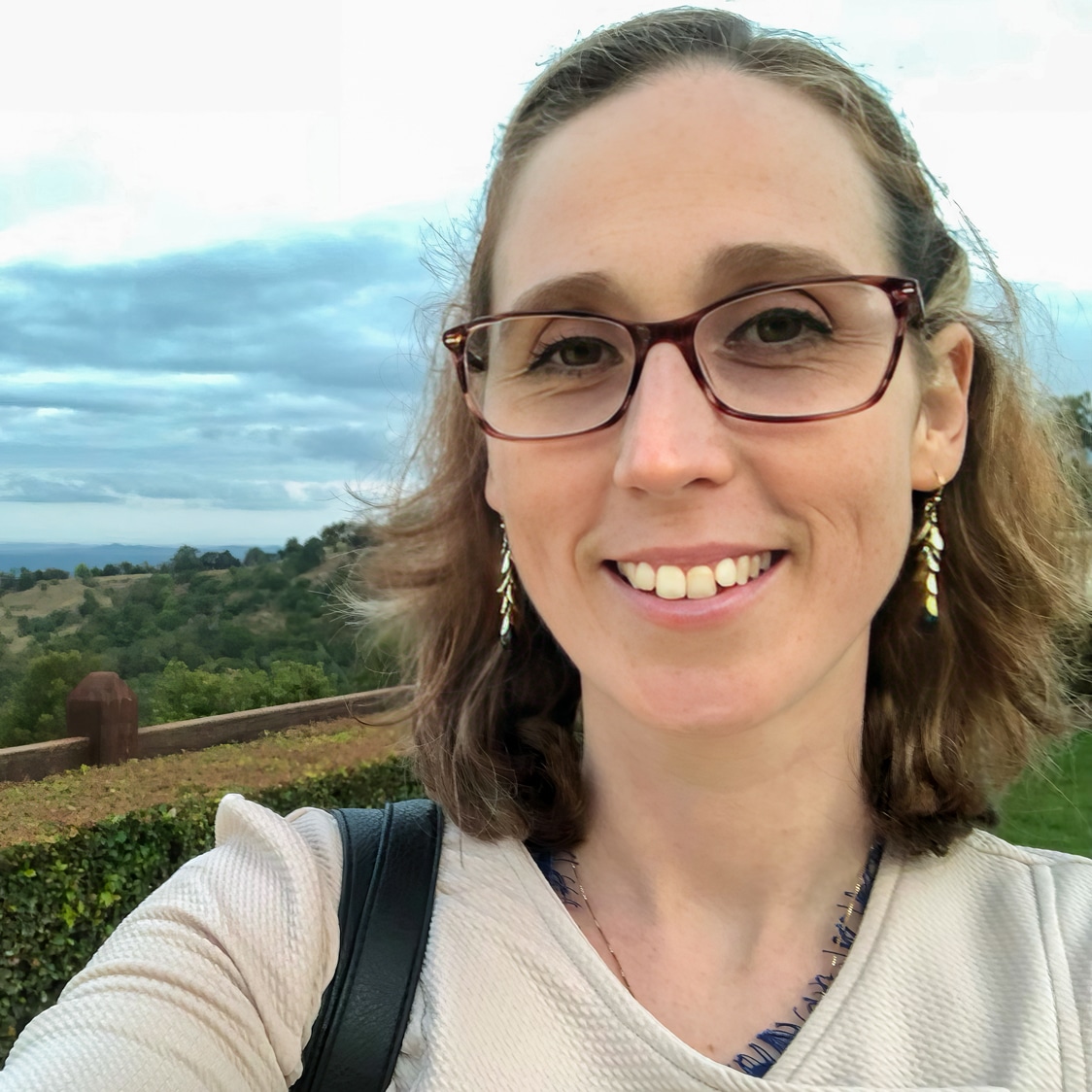 A woman with shoulder-length brown hair and glasses smiles at the camera outdoors. She is wearing dangling earrings and a beige top, with green trees and a cloudy sky in the background.