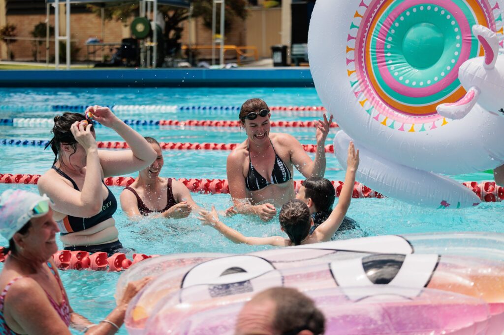A group of people enjoy swimming in a pool, playing with inflatable pool floats and laughing together on a sunny day. Red lane dividers and a colorful unicorn float are visible in the water.