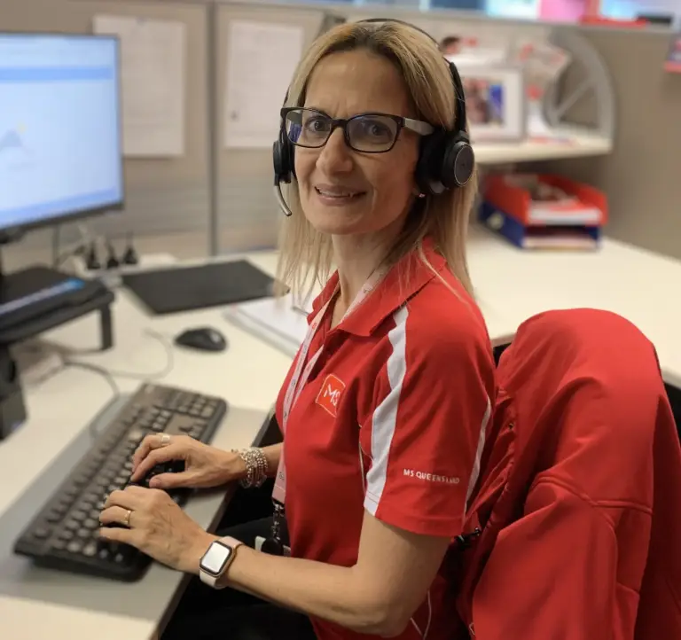 A woman wearing glasses and a headset, dressed in a red shirt, sits at an office desk typing on a keyboard. She is smiling and looking at the camera, with office supplies and a computer monitor around her.
