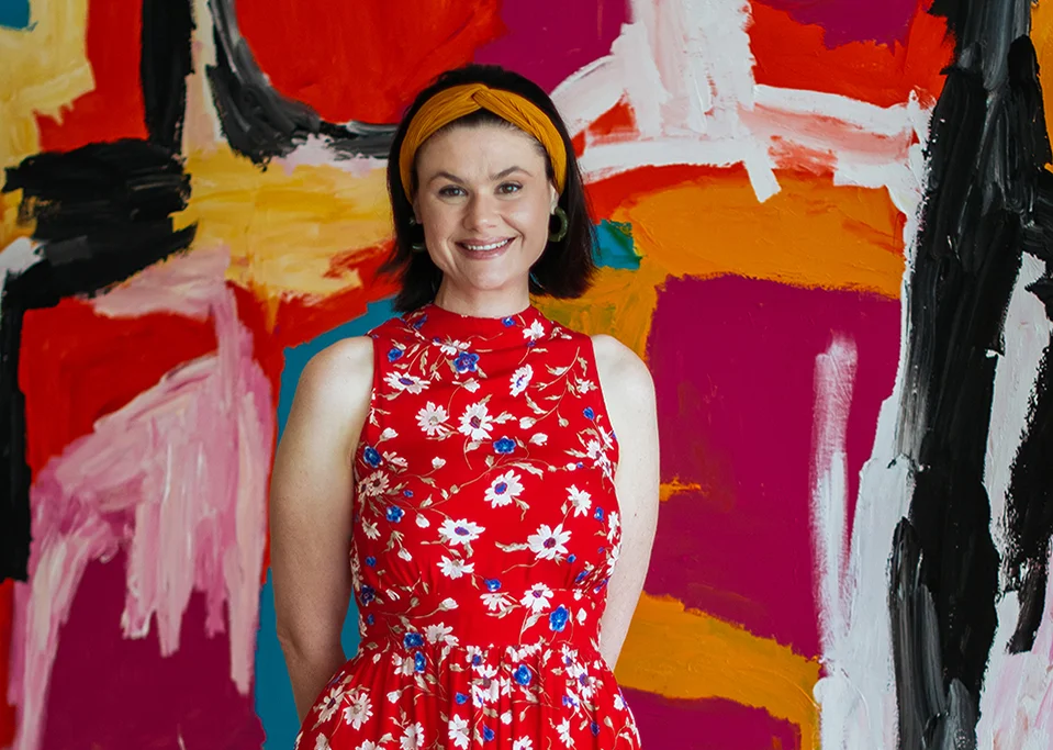 A woman in a red floral dress and an orange headband smiles while standing in front of a bright, colorful abstract painting with red, pink, black, orange, and white brushstrokes.
