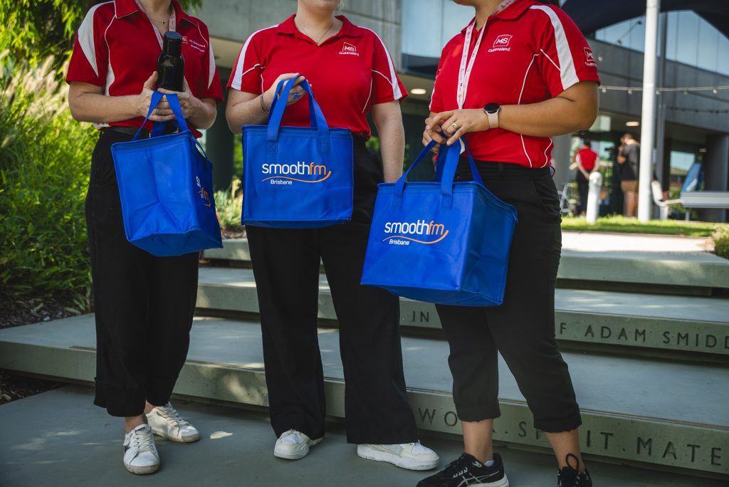 Three people in matching red shirts and black pants stand on steps outdoors, each holding a blue Smooth FM Brisbane cooler bag. Their faces are not visible.