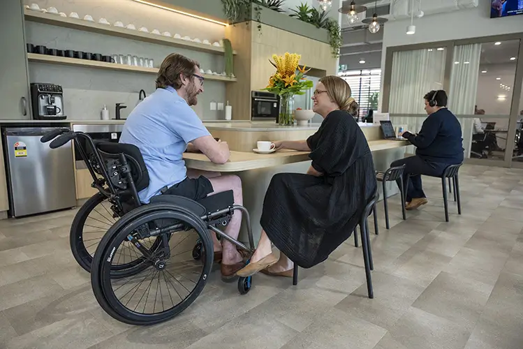 A man in a wheelchair and a woman sit at a round table in a modern, open kitchen area, having a conversation. Another person sits in the background, using a laptop. There are plants and shelves in the space.