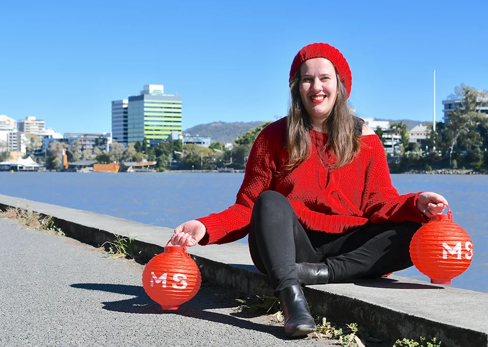 A smiling woman in a red sweater and hat sits by a riverside, holding two red lanterns with "MS" on them. City buildings and trees are visible in the background beneath a clear blue sky.