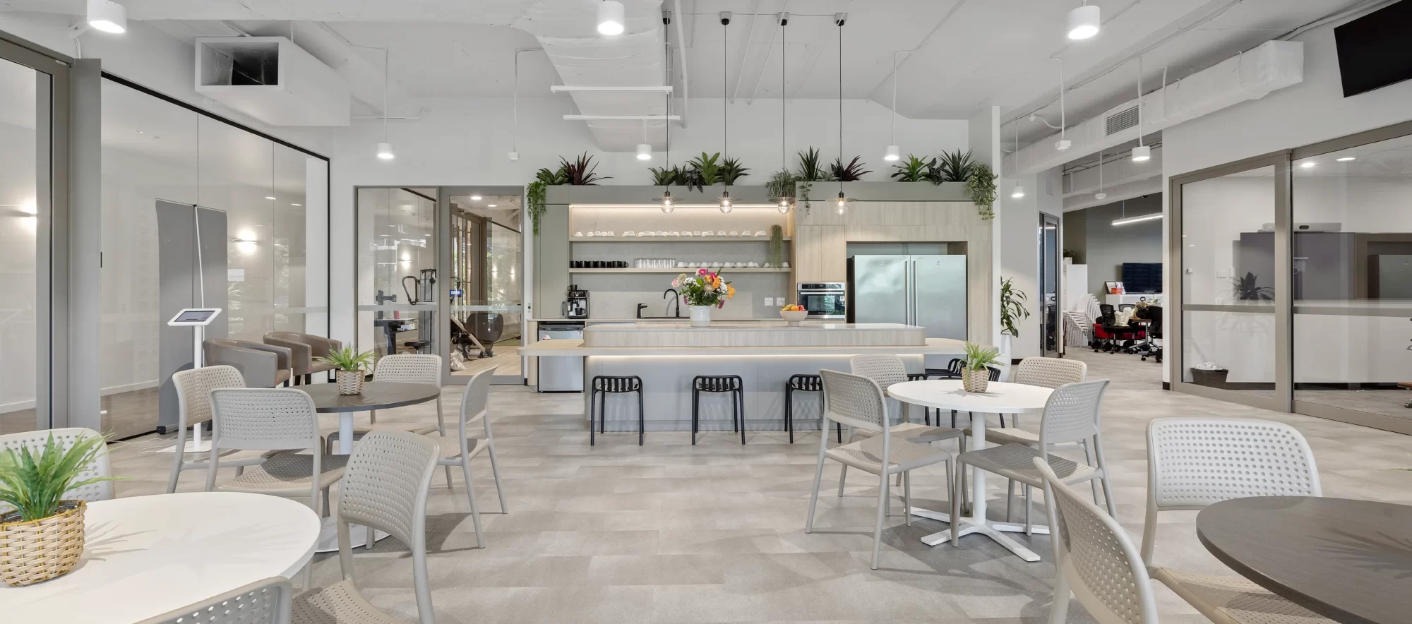 Modern office kitchen and dining area with round tables, gray chairs, a central island with barstools, hanging lights, plants, and glass-walled meeting rooms in the background. The space is bright and welcoming.