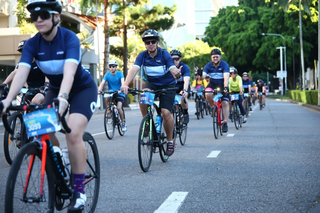 A group of cyclists wearing helmets and blue jerseys ride together on a city street during a cycling event, with trees and buildings visible in the background.