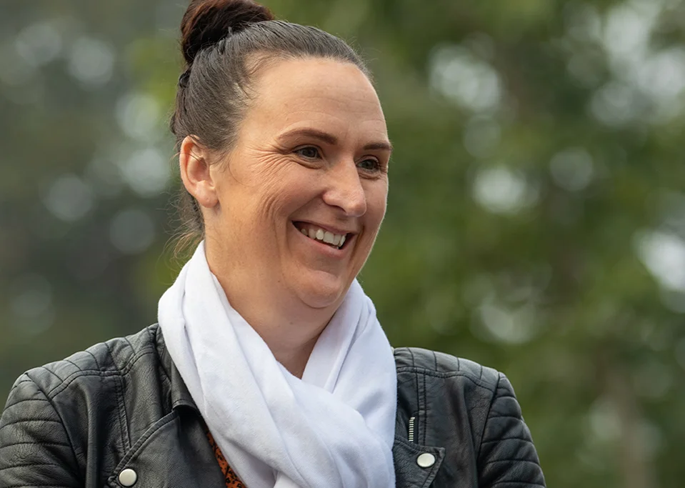 A woman with brown hair tied up is smiling outdoors. She is wearing a black leather jacket and a white scarf, with a blurred green background of trees behind her.