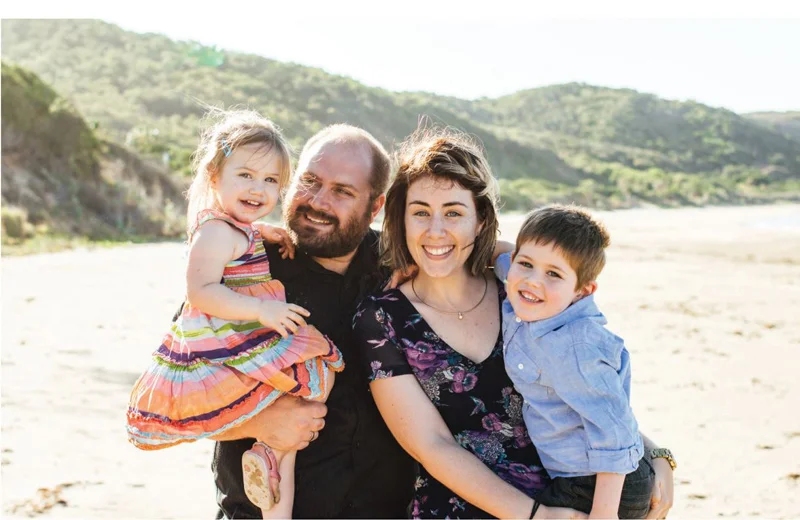 A family of four stands on a sunny beach. The father holds a smiling young girl in a colorful dress, while the mother hugs a happy young boy. Hills and greenery are in the background.