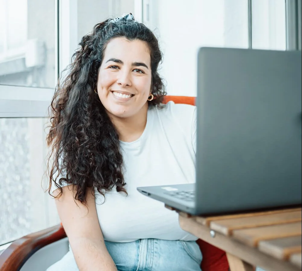 A woman with long curly hair, wearing a white shirt and jeans, sits by a window and smiles at the camera. She is seated at a wooden table with an open laptop in front of her.