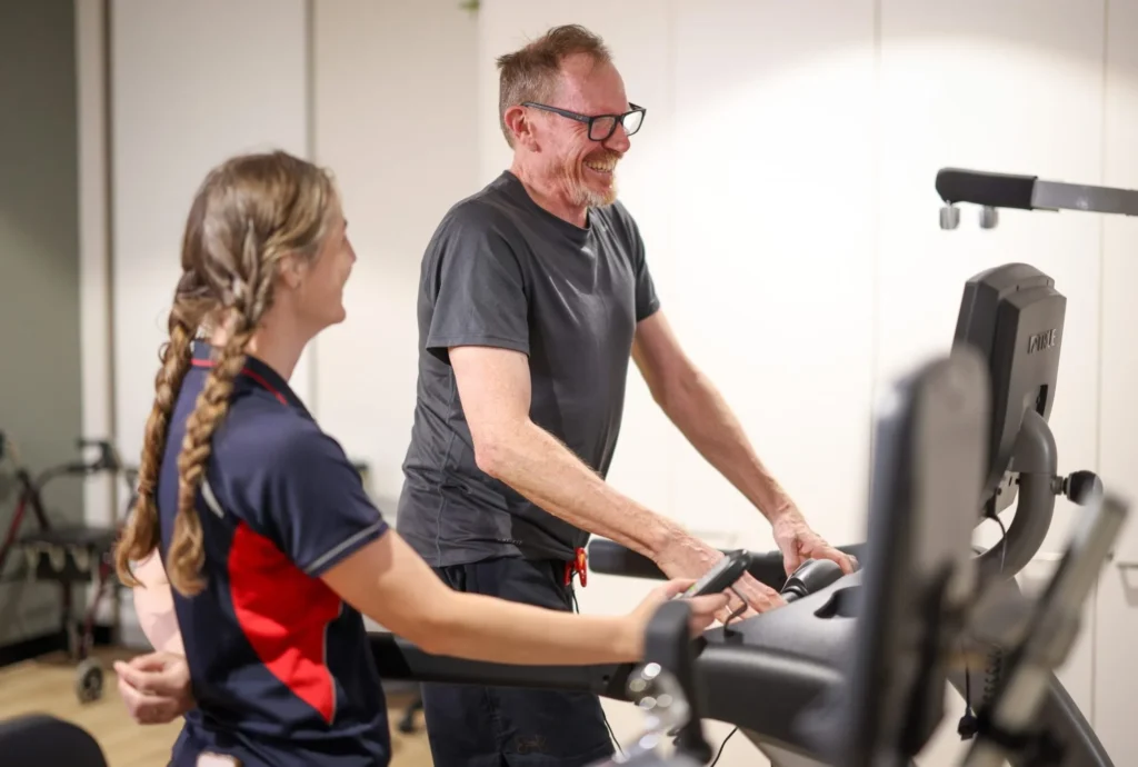 A smiling man uses a treadmill while a woman with braided hair, wearing a navy and red shirt, stands beside him, offering support and encouragement in a well-lit gym setting.
