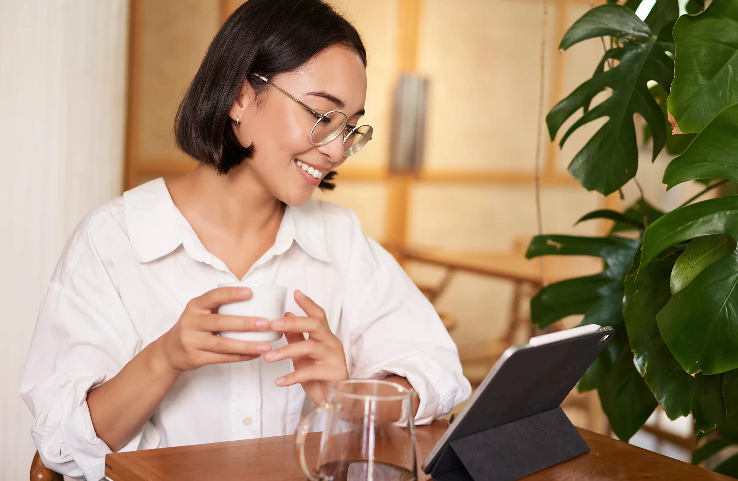 A woman with glasses and a white shirt sits at a table, smiling while holding a cup and looking at a tablet. A glass of water and a large green plant are also on the table.