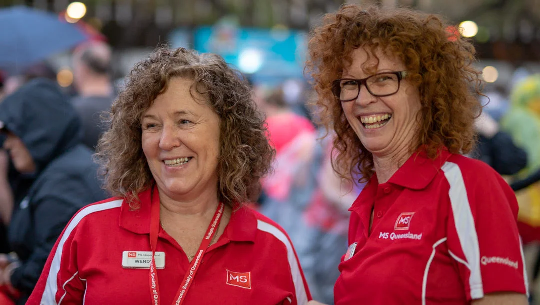 Two women wearing red MS Queensland shirts and name badges smile while standing together outdoors at an event. One woman has curly brown hair, the other has curly red hair and glasses. People and tents are visible in the background.