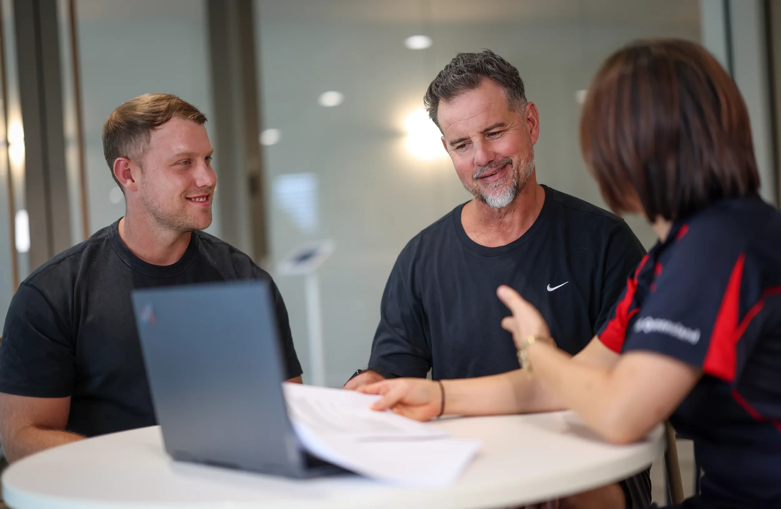Three people sit around a table with a laptop and paperwork, engaged in a friendly discussion. Two men face a woman who gestures while speaking, all appearing to be in a modern office environment.