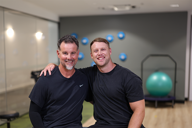 Two smiling men in athletic wear sit close together in a gym, one with his arm around the other. Exercise equipment, including a large green stability ball, is visible in the background.