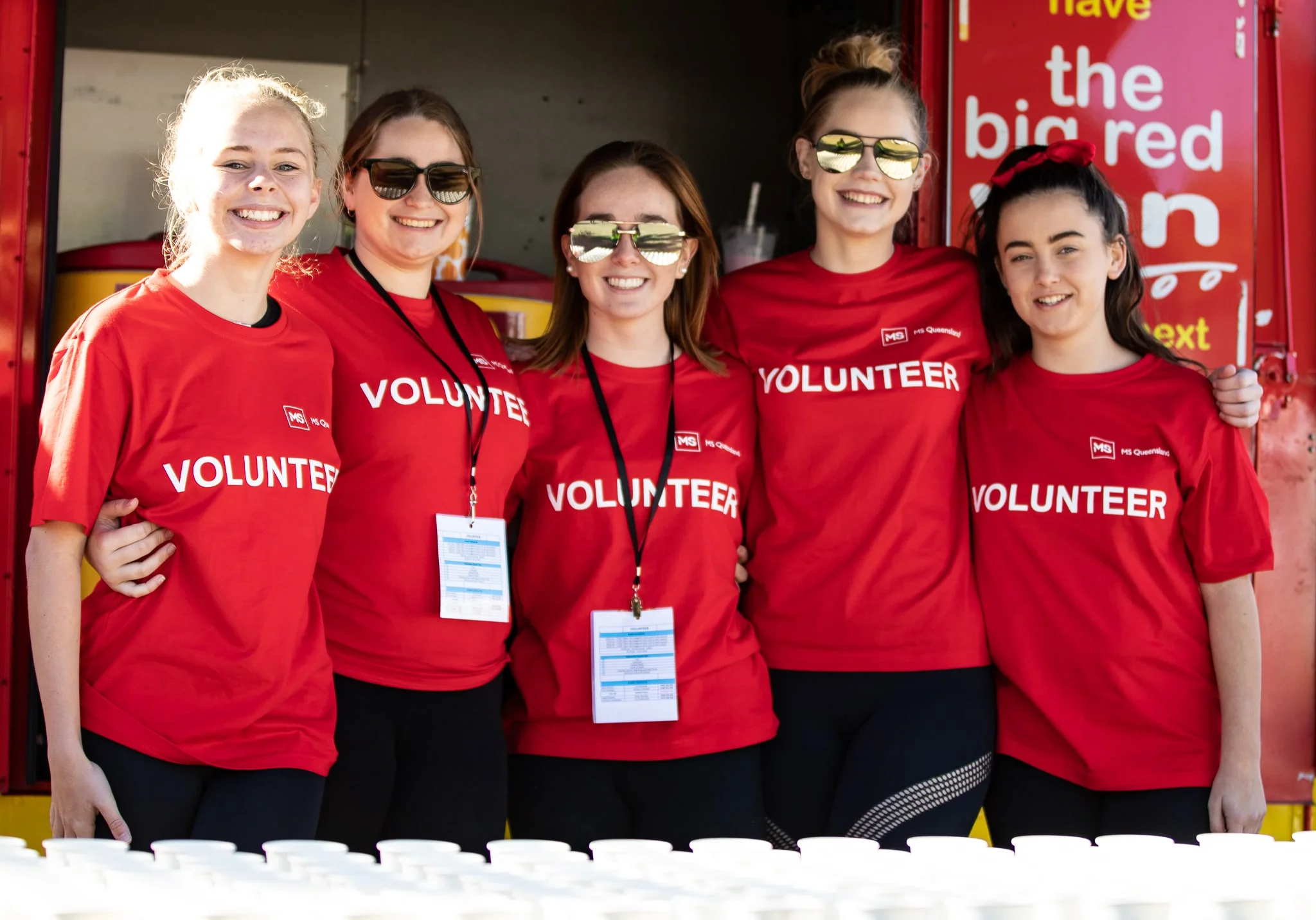 Five young women wearing matching red "VOLUNTEER" shirts stand side by side, smiling at the camera. They are outdoors, and white cups are lined up in the foreground.