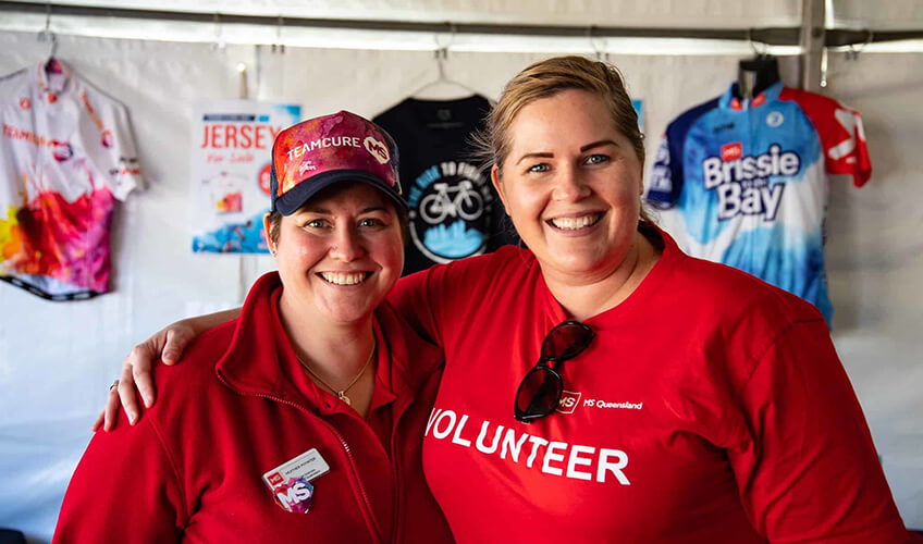 Two smiling women wearing red volunteer shirts and caps stand together inside a tent. Cycling jerseys hang on the wall behind them. One woman has her arm around the other’s shoulder.