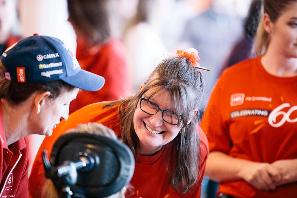 A woman in glasses and an orange shirt smiles warmly at a person in a blue cap, with others in red shirts blurred in the background. The setting appears lively and cheerful.