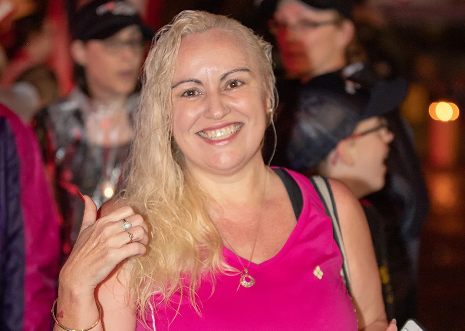 A smiling woman with long blonde hair wearing a bright pink sleeveless top stands in a crowd at night, giving a thumbs up. She is wearing jewelry and other people are visible in the blurred background.