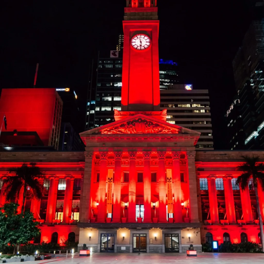 A historic building with columns and a clock tower is illuminated with bright red lights at night, standing out against the dark sky and surrounding modern buildings.