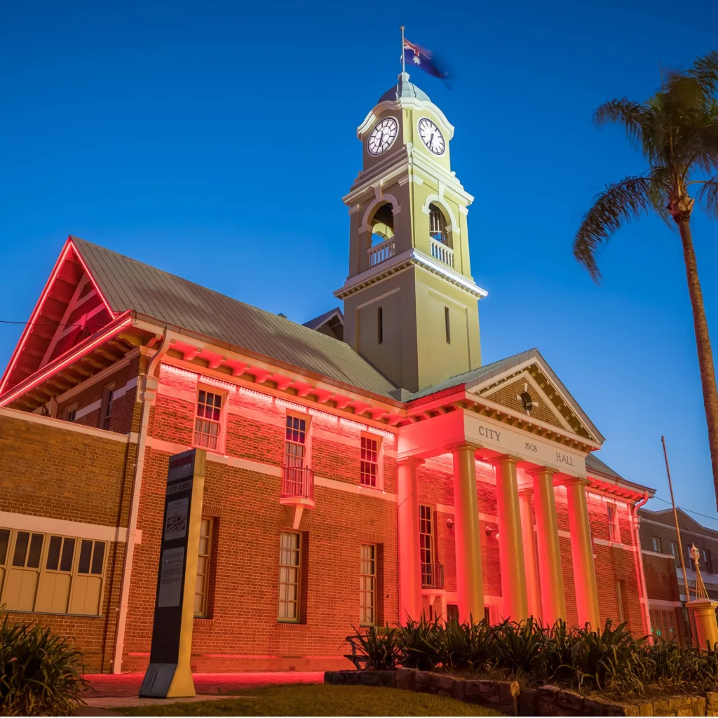Historic brick city hall building illuminated with red and yellow lights at dusk, featuring a tall clock tower with a flag on top, white columns at the entrance, and a palm tree on the right.