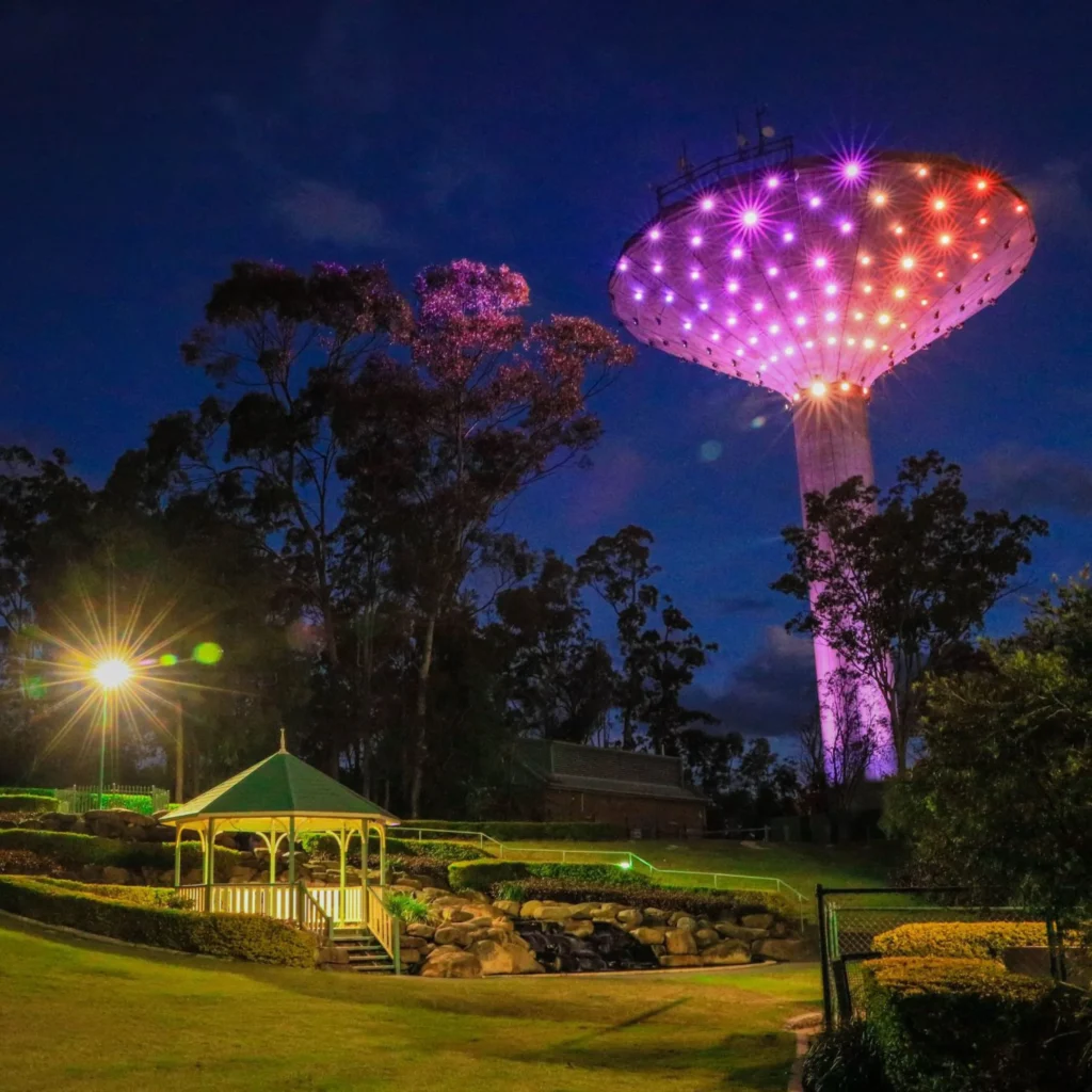 A large water tower lit with purple and orange lights stands behind tall trees at dusk. In the foreground, a small white gazebo sits on a grassy lawn near a rocky garden.