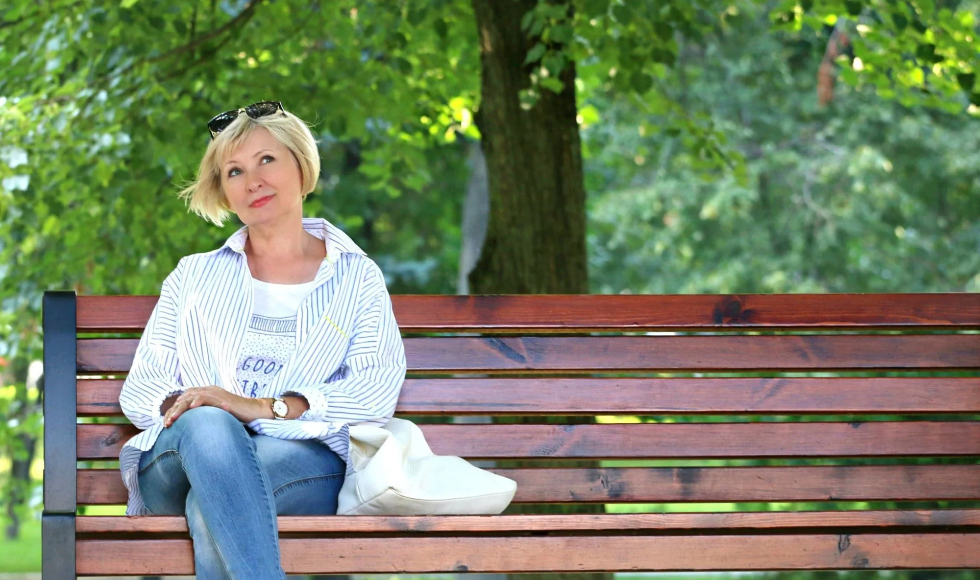 A woman with short blonde hair sits on a wooden park bench, looking up thoughtfully. She is wearing jeans, a striped shirt, sunglasses on her head, and a white bag sits beside her. Green trees fill the background.
