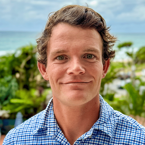 A man with short, wavy brown hair and blue eyes smiles at the camera. He is wearing a blue and white checkered shirt. Lush green plants and a cloudy sky are visible in the background.