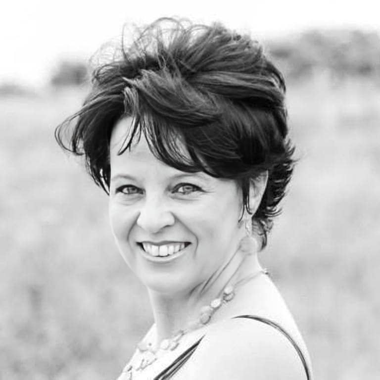 Black and white photo of a woman with short, dark, styled hair, smiling at the camera. She is outdoors with a blurred background and is wearing a sleeveless top and a beaded necklace.