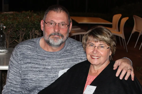 A man with glasses and a gray sweater has his arm around a smiling woman wearing glasses and a dark shawl. They are seated together at a table, both wearing name tags. Indoor setting with chairs and tables in the background.