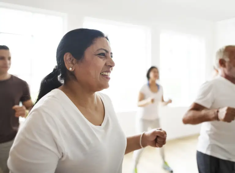 A group of adults in casual workout clothes smile and exercise together in a bright, sunlit room. The focus is on a woman in the foreground, looking happy and energized.
