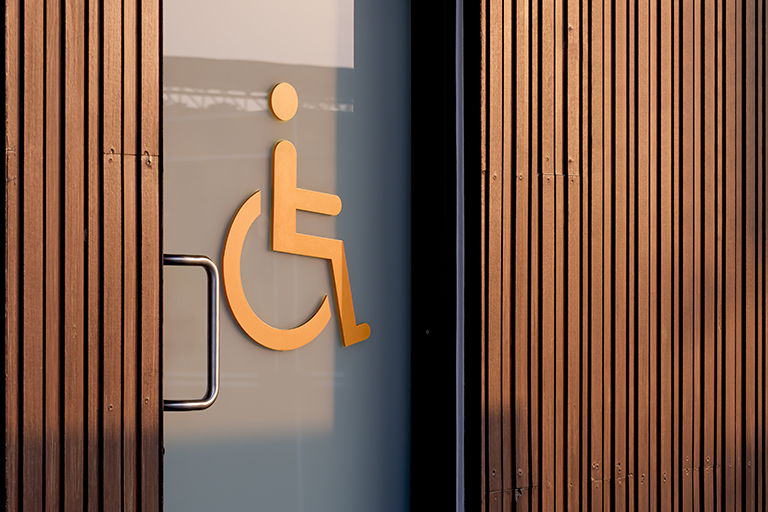 A glass door with a yellow wheelchair accessible symbol, set in a wall with vertical wooden panels. The metal door handle is visible on the left side of the door.