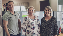 Three people stand indoors, smiling at the camera. They are in an office setting with cubicles and papers pinned to the walls behind them. The group consists of two women and one man.