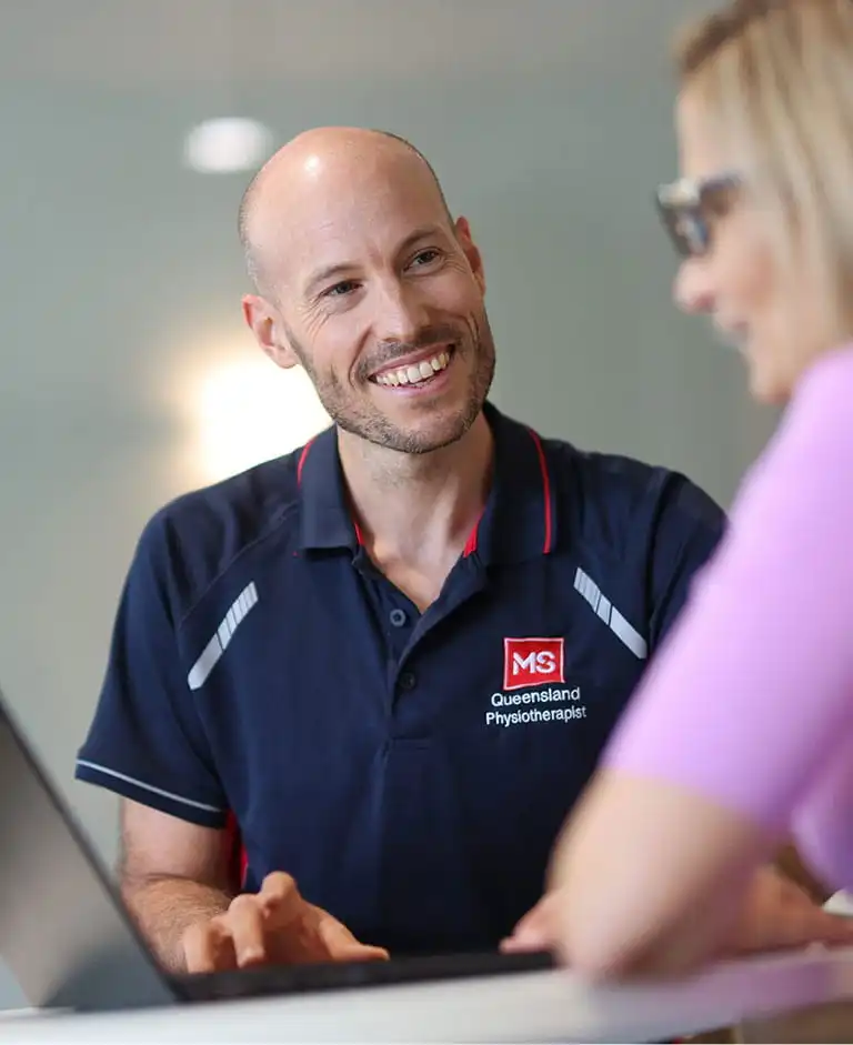 A smiling man in a "Queensland MS Physiotherapist" shirt talks to a woman across a table, both looking at a laptop. The setting appears to be a professional or medical office.