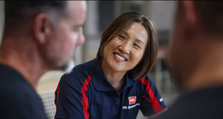 A woman wearing a navy blue shirt with red accents and an MS Queensland logo smiles while talking with two men in a casual indoor setting.
