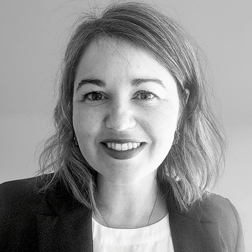 A woman with shoulder-length wavy hair smiles at the camera. She is wearing a dark blazer over a light top, and the photo is in black and white.