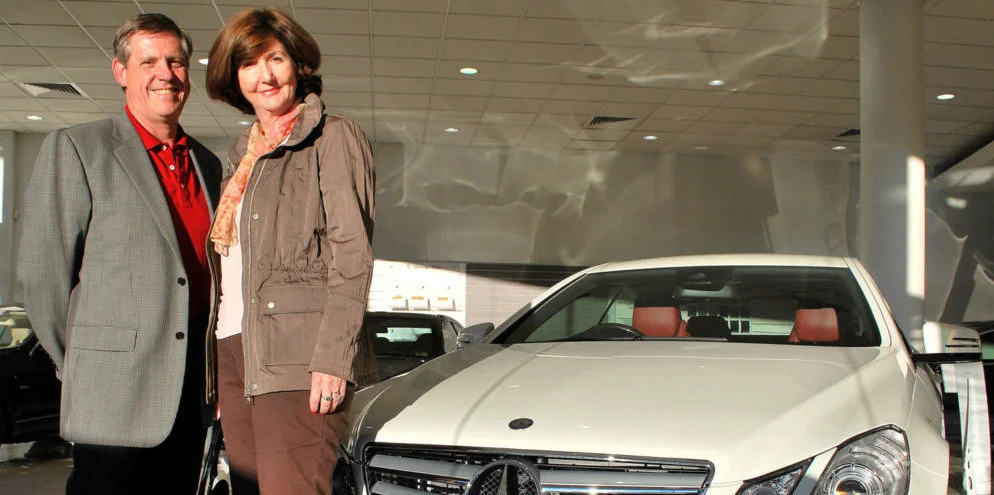 A man and woman stand smiling next to a white Mercedes-Benz car inside a bright showroom with large windows and overhead lights.
