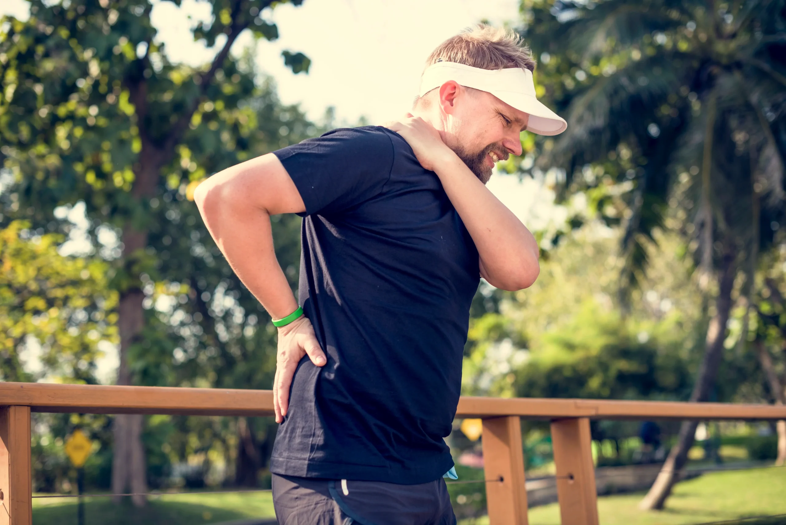 A man in athletic attire and a visor stands outdoors, holding his lower back and neck with a pained expression, suggesting discomfort or injury. Trees and greenery are visible in the background.