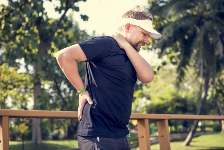 A man wearing a white visor and black shirt stands outdoors, holding his lower back and neck with a pained expression, suggesting discomfort or injury, surrounded by trees and sunlight.