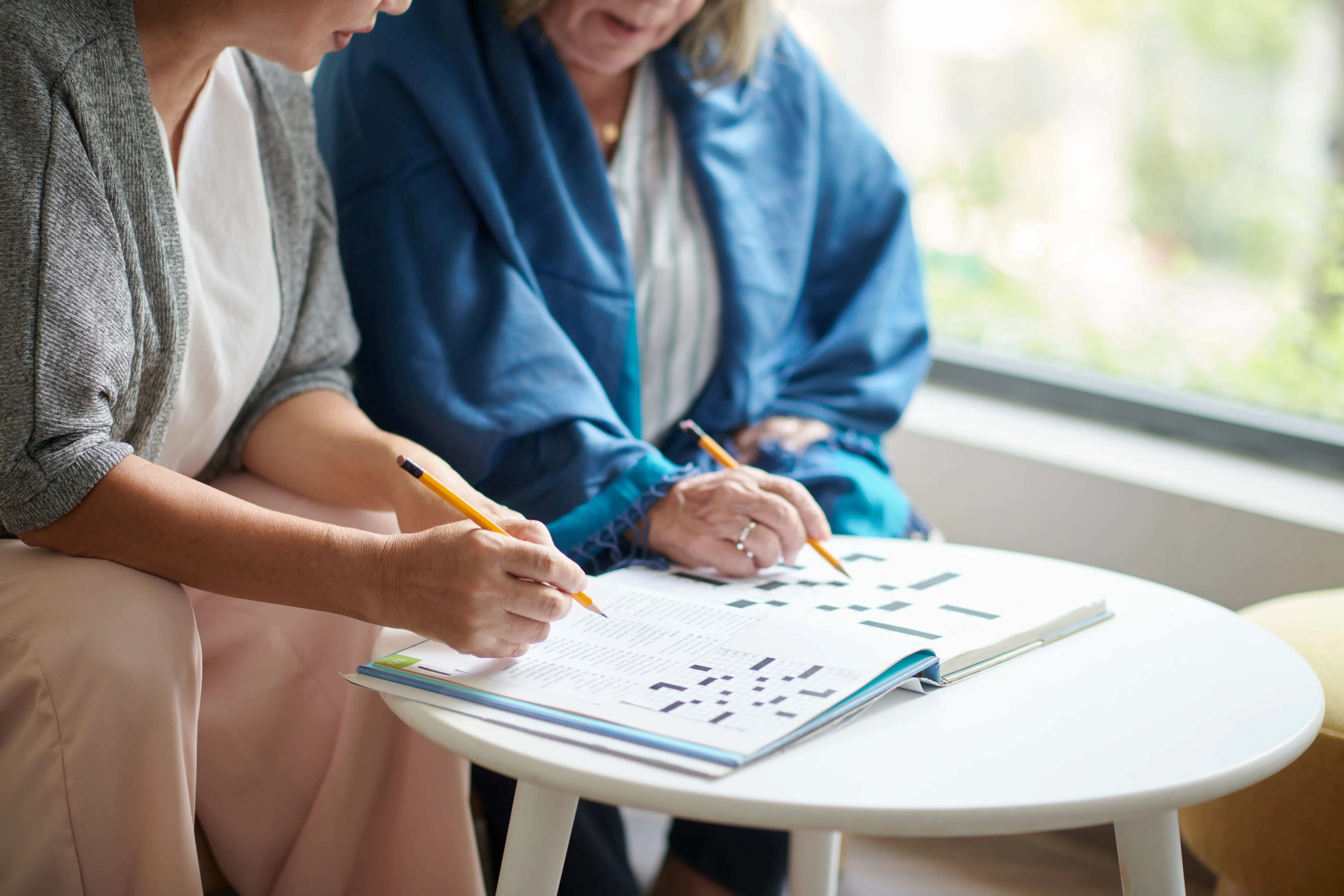 Two women sitting at a round white table by a window, working together on crossword puzzles in a book, each holding a pencil and focusing on the pages.