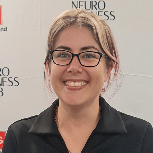A smiling person with light hair, wearing glasses and a black collared shirt, stands in front of a white backdrop with partially visible text that reads "NEURO BUSINESS LAB.
