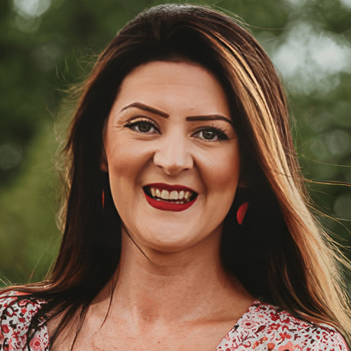 A woman with long dark hair and red lipstick smiles outdoors, wearing a floral top and red earrings, with a blurred green background.
