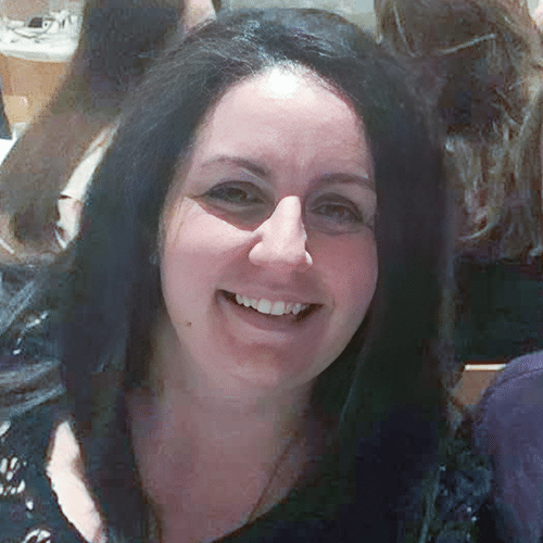 A woman with long dark hair and a black top smiles at the camera. There are other people blurred in the background.