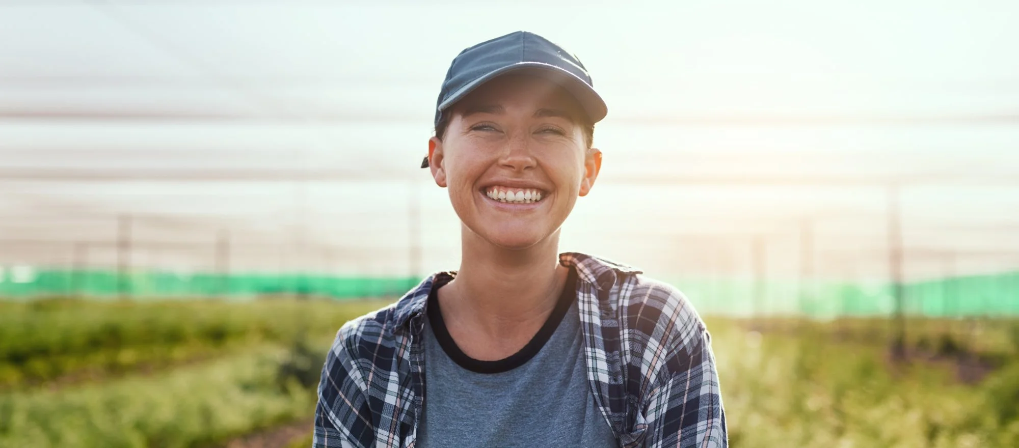 A smiling person wearing a baseball cap and plaid shirt stands outdoors in a sunlit field or greenhouse, with green plants and netting visible in the background.