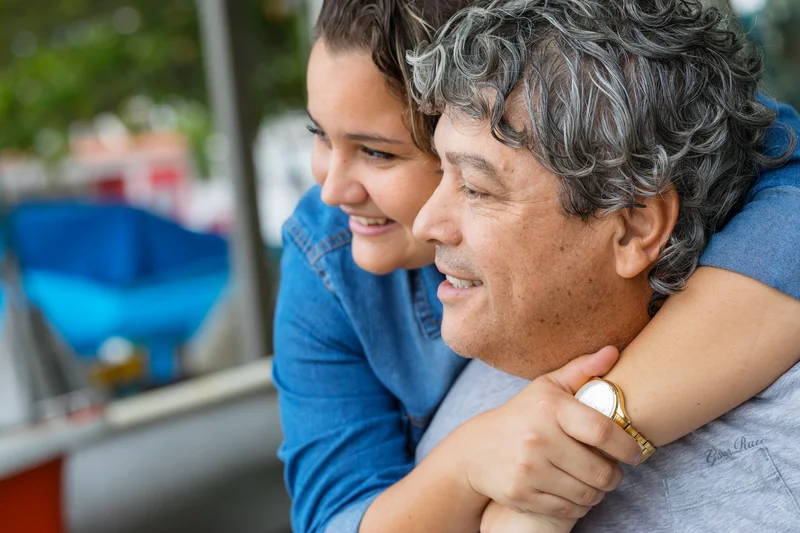 A smiling young woman hugs an older man from behind. Both appear happy and are looking into the distance. The background is blurred, showing some greenery and outdoor elements.