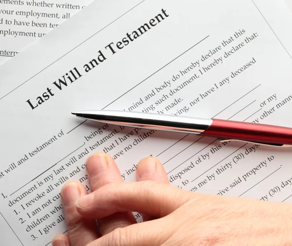 A hand holding a pen hovers over a document titled "Last Will and Testament," ready to sign. The paper contains printed legal text and signature lines.
