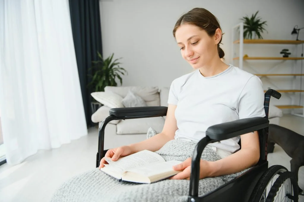 A young woman sits in a wheelchair indoors, holding and reading a book on her lap. She is wearing a white t-shirt and light blanket, and appears calm and content in a bright, modern living room.