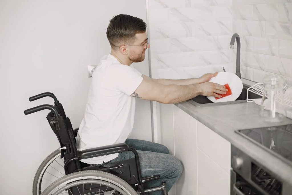 A man in a wheelchair is washing dishes at a kitchen sink, using a red sponge to clean a plate. The kitchen has a modern design with white and gray tones.