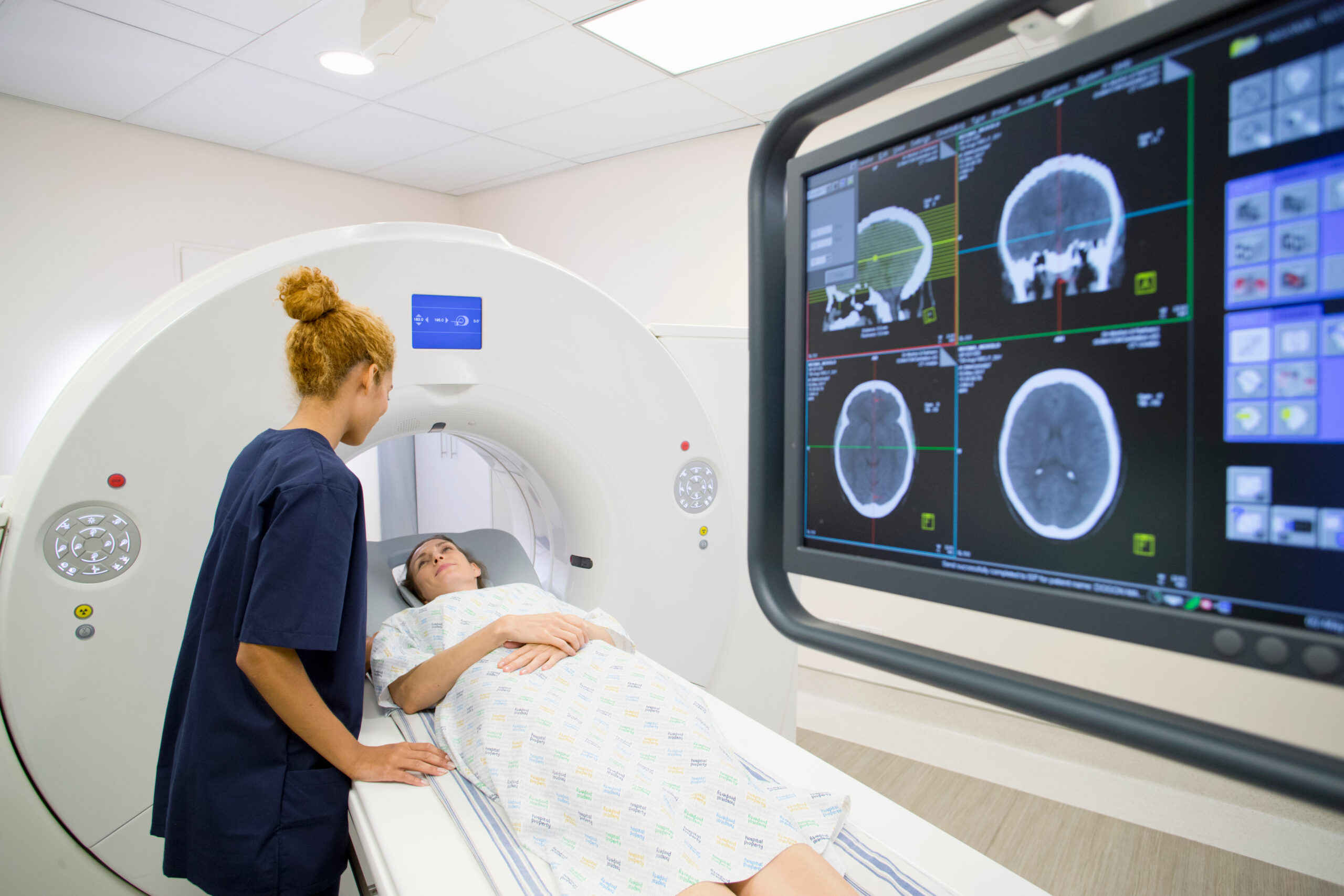 A patient lies on a CT scanner bed while a healthcare worker stands beside them. A monitor displays brain scan images in the foreground. The setting appears to be a modern medical imaging room.