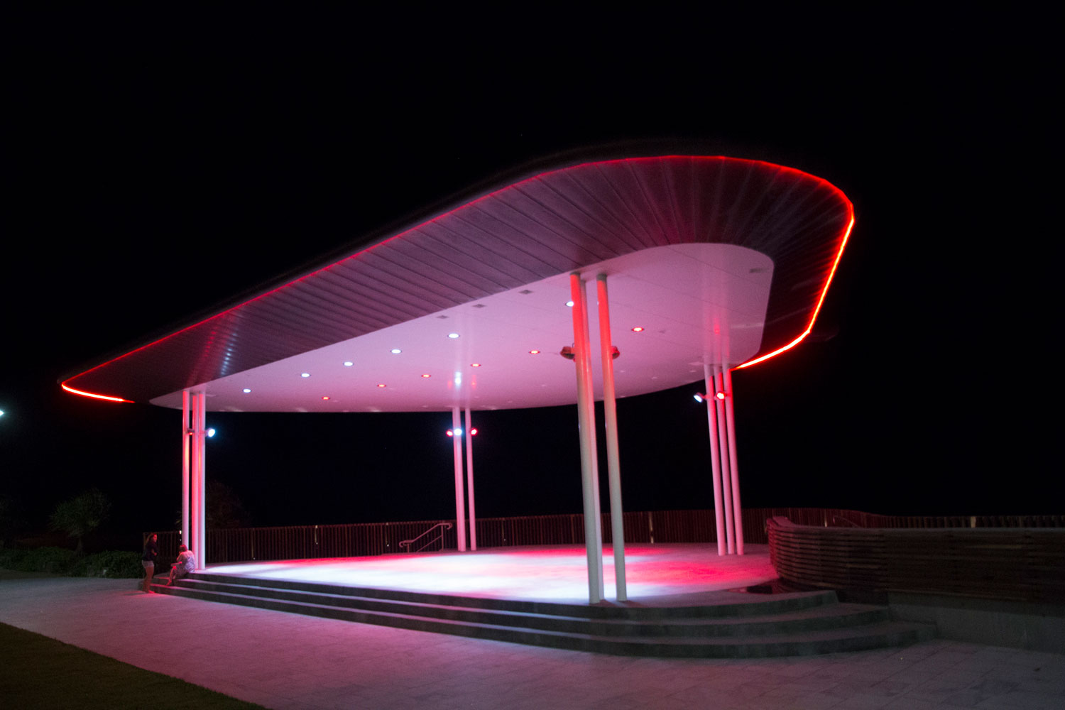 Modern outdoor stage with a curved roof is illuminated by red and white lights at night, casting a bright glow on the ground. The area appears empty except for a person sitting at the edge.